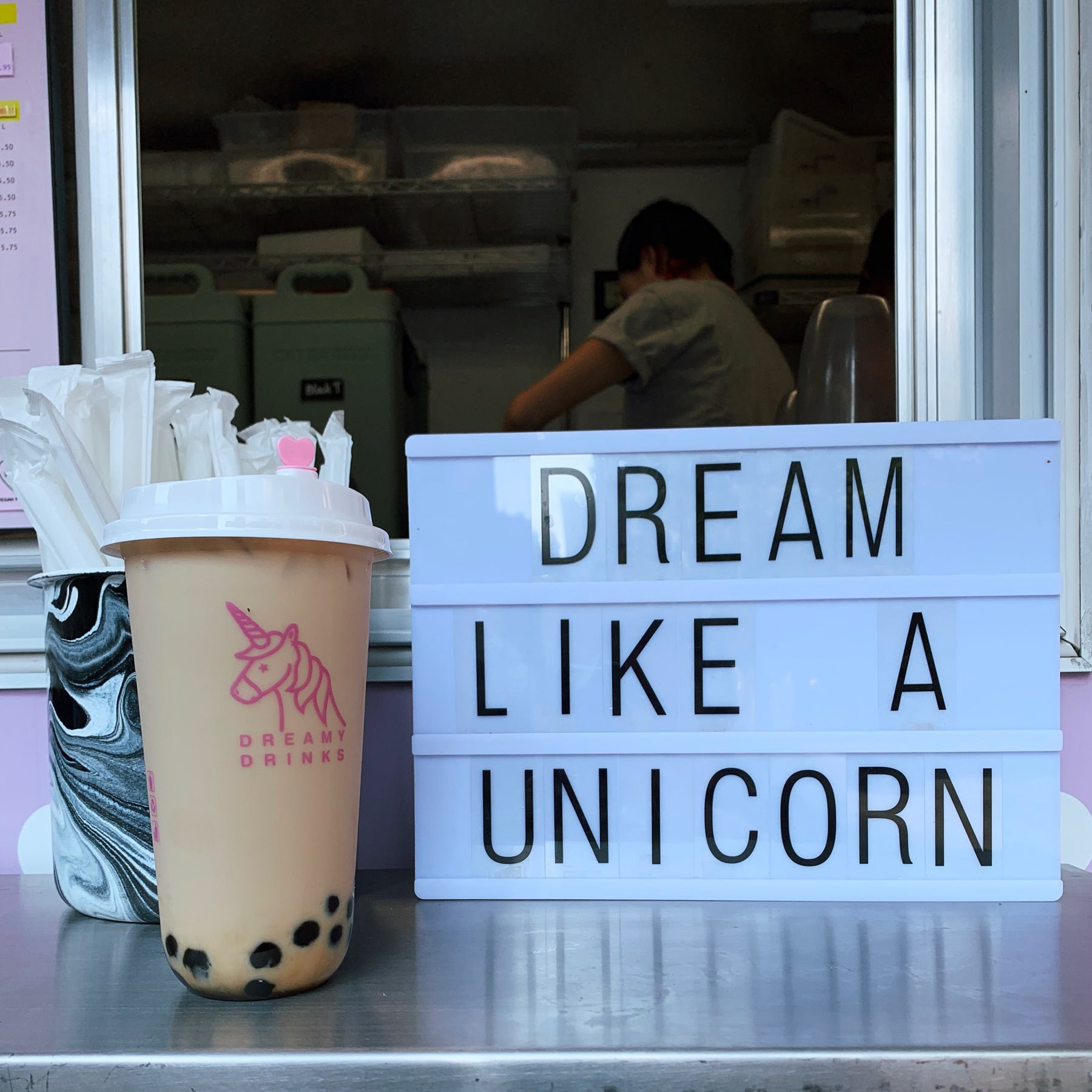 A food truck. A cup of bubble tea is on a shelf next to a aign that says, "Dream like a unicorn."
